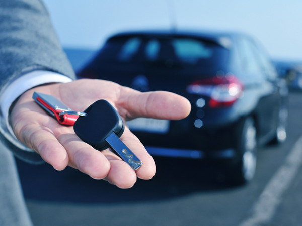 man in suit offering a car key to the observer, with a car in the background